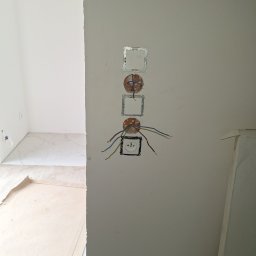 Adam Remontuje - Ściana z przygotowanymi otworami i przewodami pod gniazdka elektryczne i włączniki, widok na wykończoną podłogę w drugim pomieszczeniu. Prace instalacyjne w trakcie remontu.