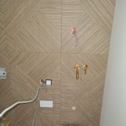 Adam Remontuje - Wzorzysta, beżowa ściana z płytek w łazience, z widocznymi instalacjami elektrycznymi i hydraulicznymi przed montażem armatury. Uwagę przykuwa geometryczny wzór płytek.