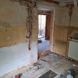 Adam Remontuje - Wnętrze w trakcie remontu: odsłonięta cegła, kable, narzędzia, usunięte fragmenty podłogi. Widoczne ślady starej tapety i instalacji elektrycznej.