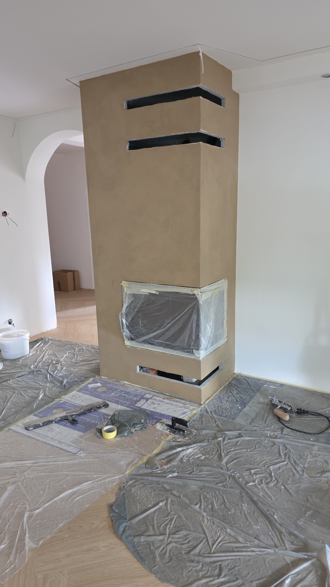 Wnętrze w trakcie prac: obudowa kominka z otworami wentylacyjnymi, zabezpieczona folią, narzędzia na podłodze, widok na łukowe przejście do innego pomieszczenia.