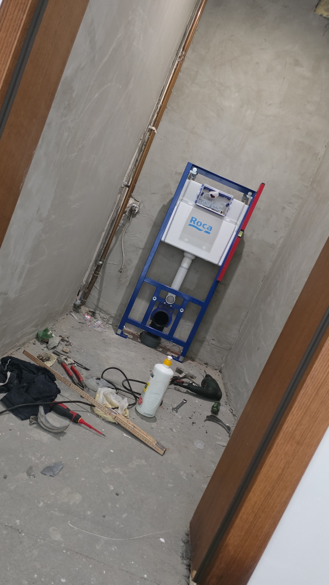 Montaż stelaża podtynkowego WC Roca w surowej łazience. Widoczne narzędzia i materiały budowlane na podłodze, miedziane rury na ścianie.