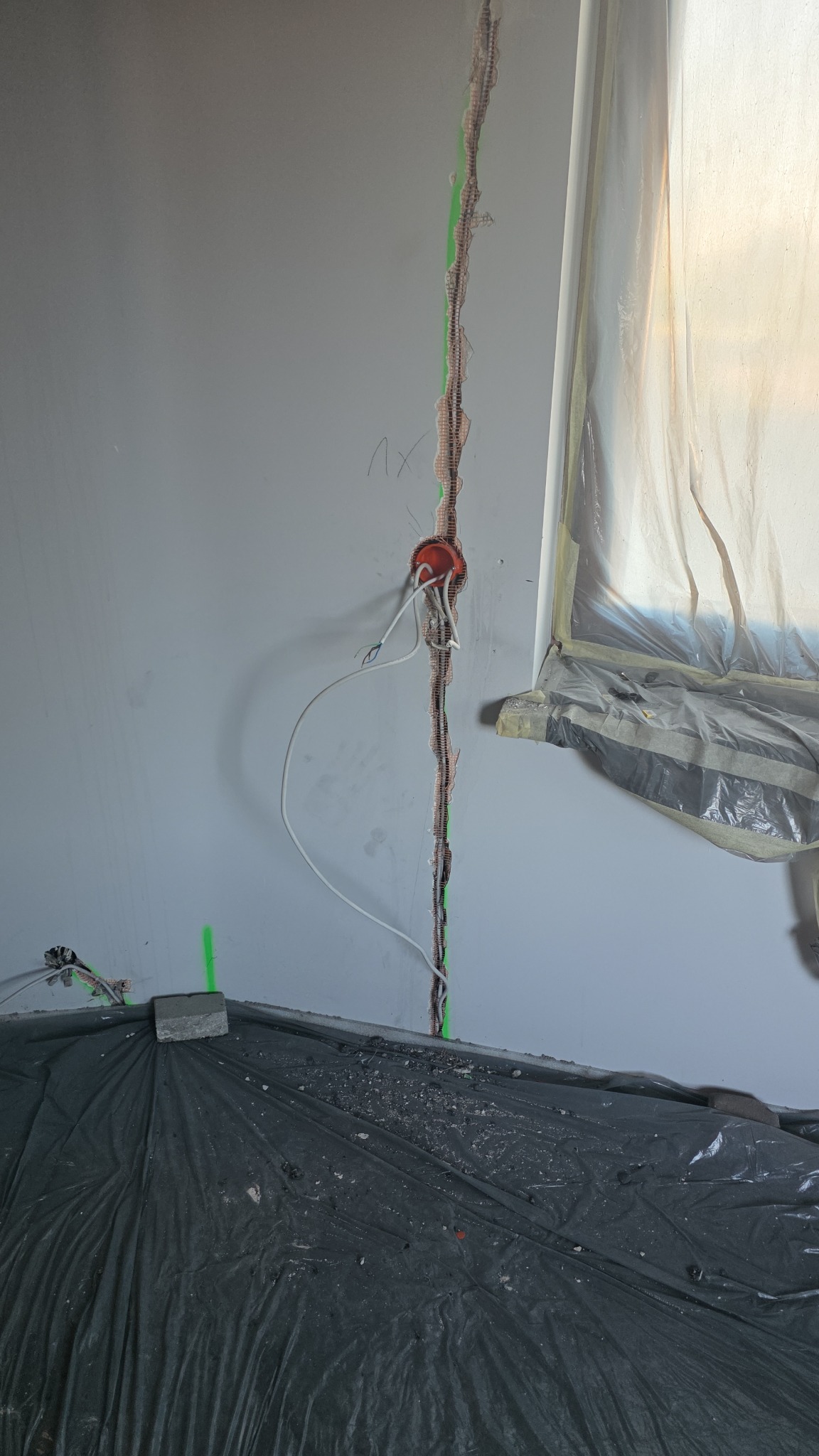 Bruzda w ścianie z kablem elektrycznym i puszką instalacyjną, zabezpieczone okno folią malarską, podłoga zakryta czarną folią ochronną. Prace instalacyjne w trakcie remontu.