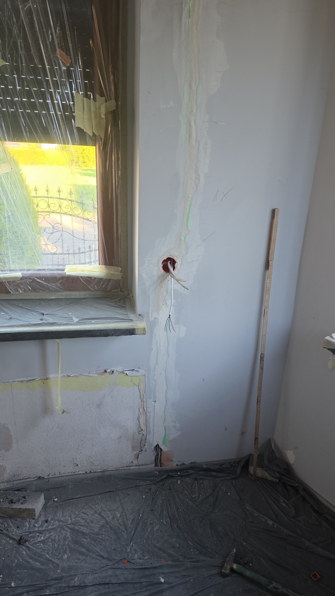Ściana w trakcie szpachlowania z widocznymi przewodami elektrycznymi i zabezpieczonym oknem folią. Na podłodze folia ochronna, widoczna łata i poziomica.
