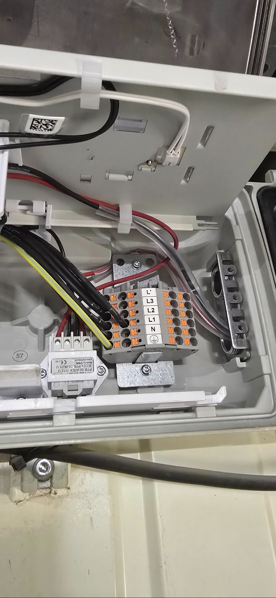 Otwarta skrzynka elektryczna z widocznymi przewodami, złączkami i elementami montażowymi. Widoczne oznaczenia L1, L2, L3, N, oraz symbol uziemienia.