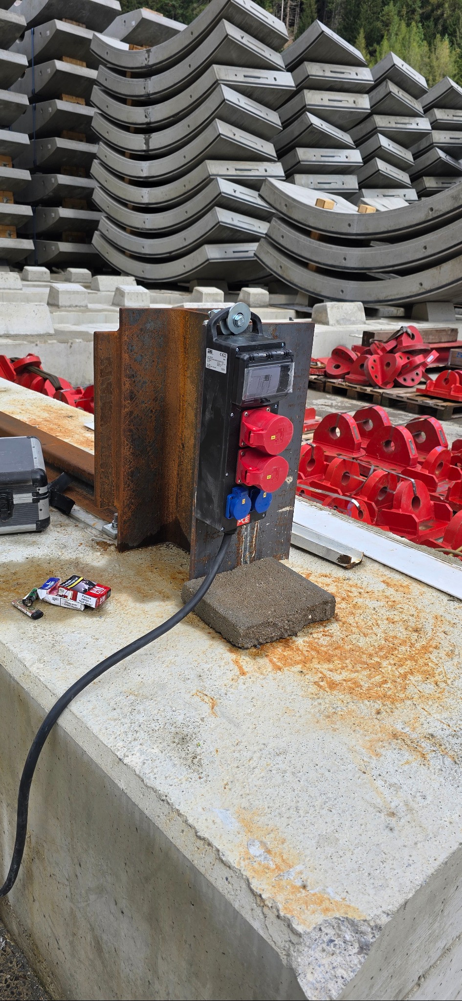 Tymczasowa skrzynka elektryczna z czerwono-niebieskimi gniazdami na betonowym fundamencie, w tle stos prefabrykatów i czerwone elementy montażowe.