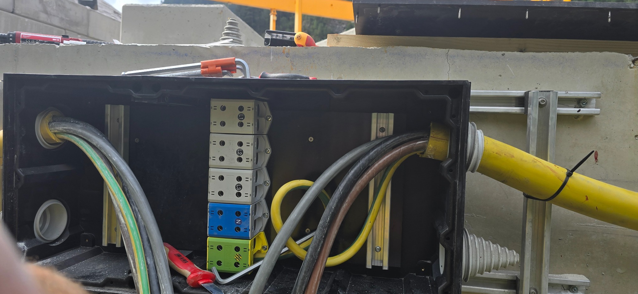 Czarna skrzynka elektryczna z kolorowymi złączkami i kablami w żółtej osłonie, zamontowana na szarej ścianie z metalowymi szynami. Widoczne narzędzia.