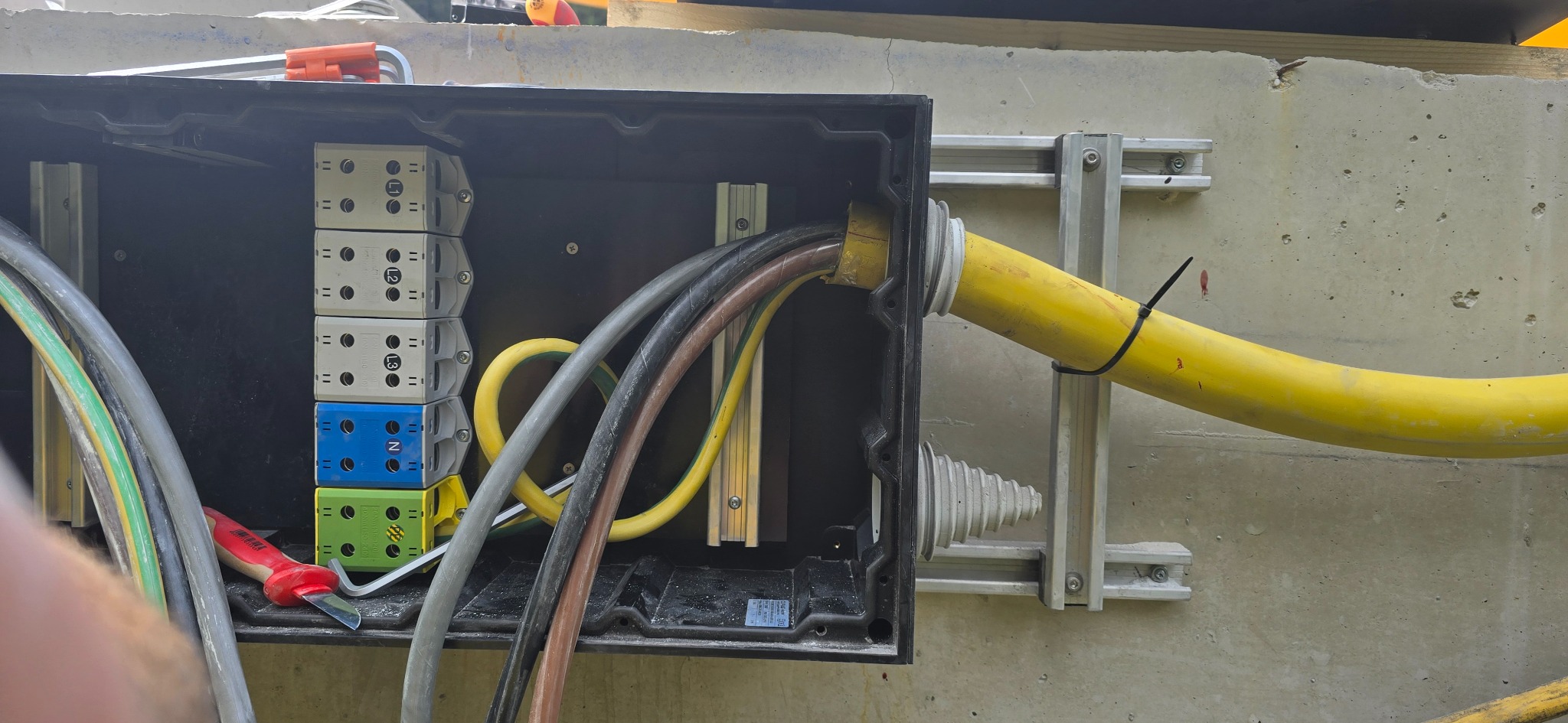 Czarna skrzynka elektryczna z podłączonymi przewodami i listwami zaciskowymi na betonowej ścianie. Widoczny żółty peszel i narzędzie.