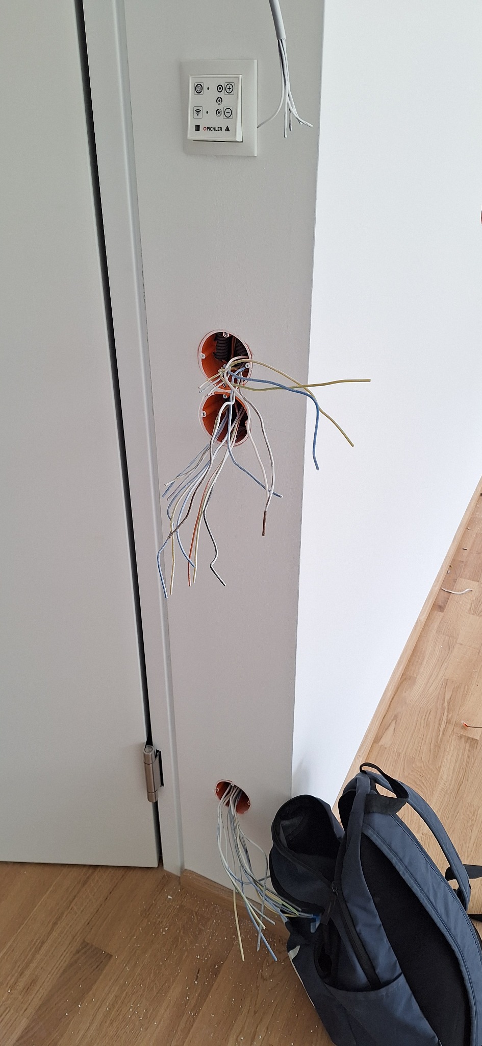 Przygotowanie do montażu gniazdek elektrycznych w ścianie obok białych drzwi. Widoczne kable, puszki instalacyjne i panel sterowania oświetleniem.