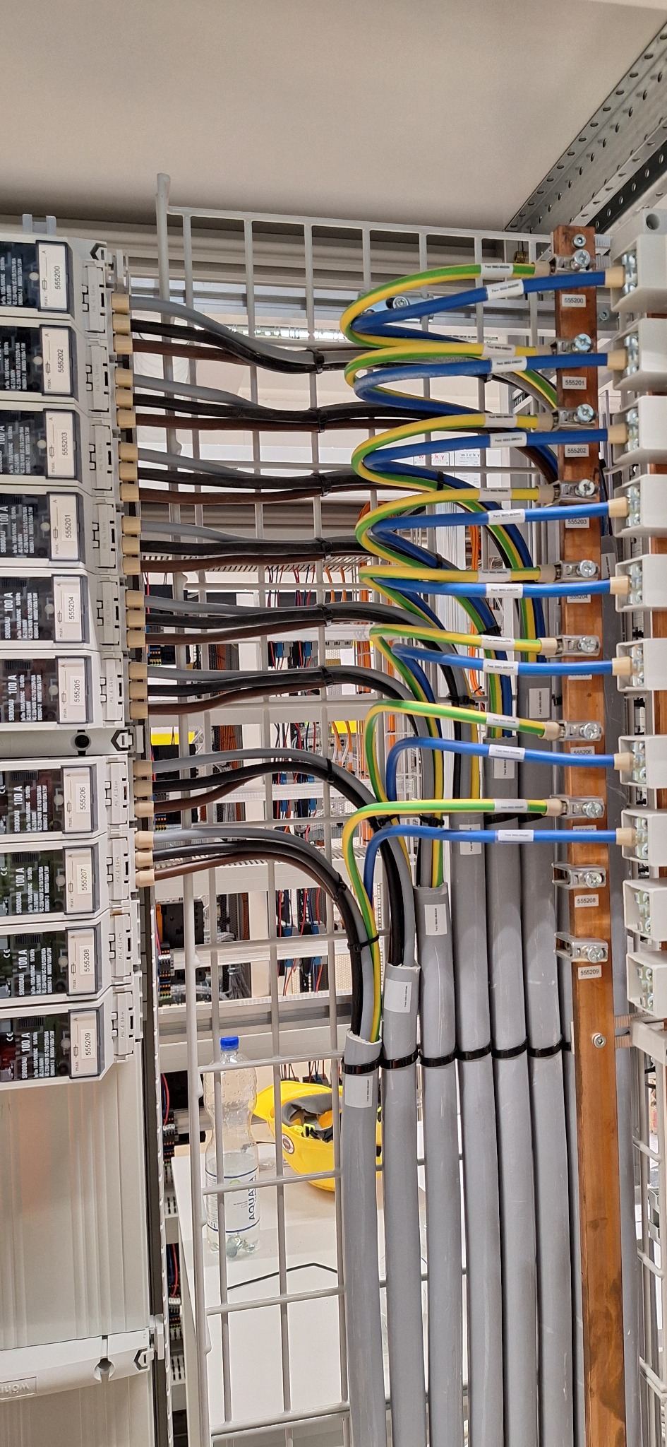 Rozdzielnica elektryczna z precyzyjnie ułożonymi kablami oznaczonymi etykietami, osadzonymi w metalowej siatce. Widoczne zabezpieczenia i elementy montażowe.