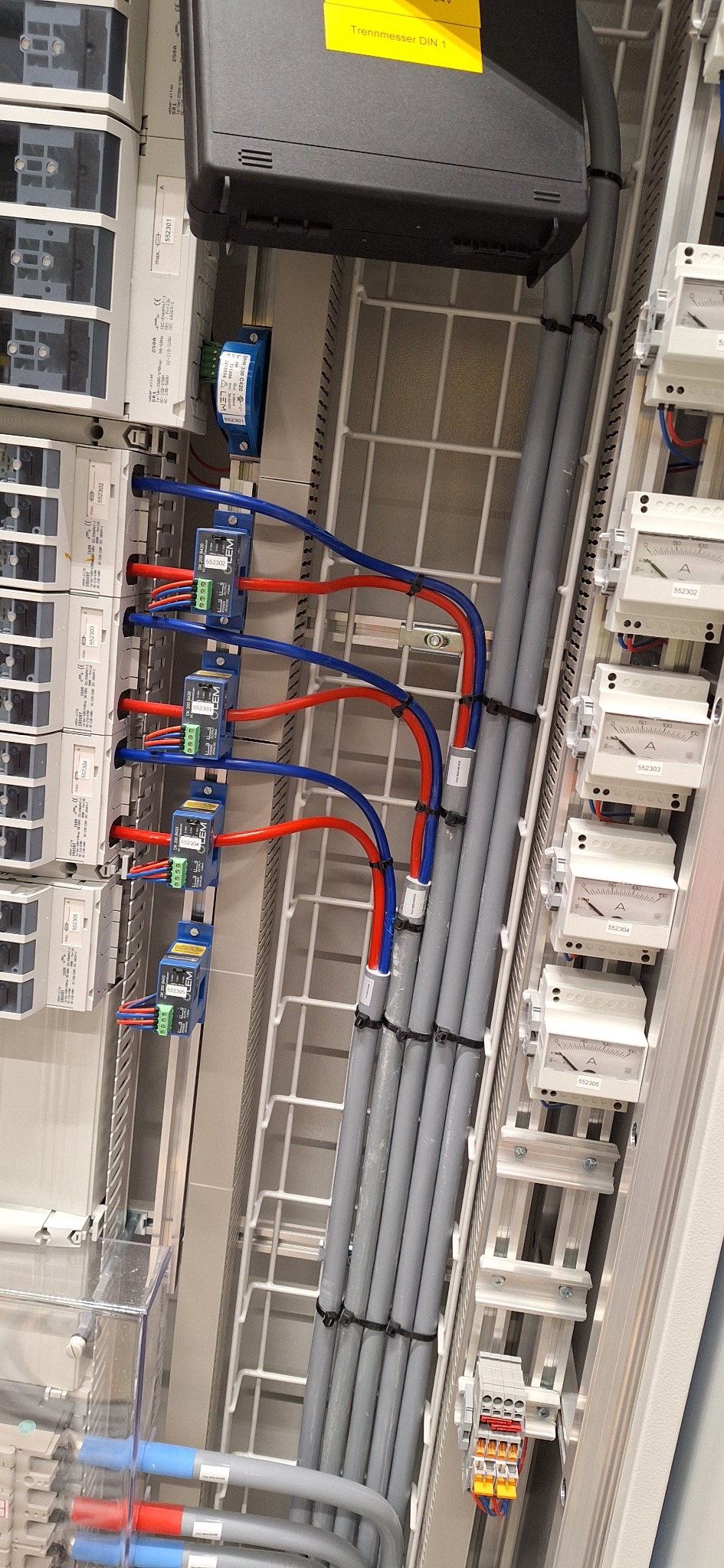 Panel elektryczny z przewodami w kolorach niebieskim i czerwonym, szare rury kablowe, biała kratka. Widoczne liczniki i moduły. Instalacja elektryczna w rozdzielnicy.