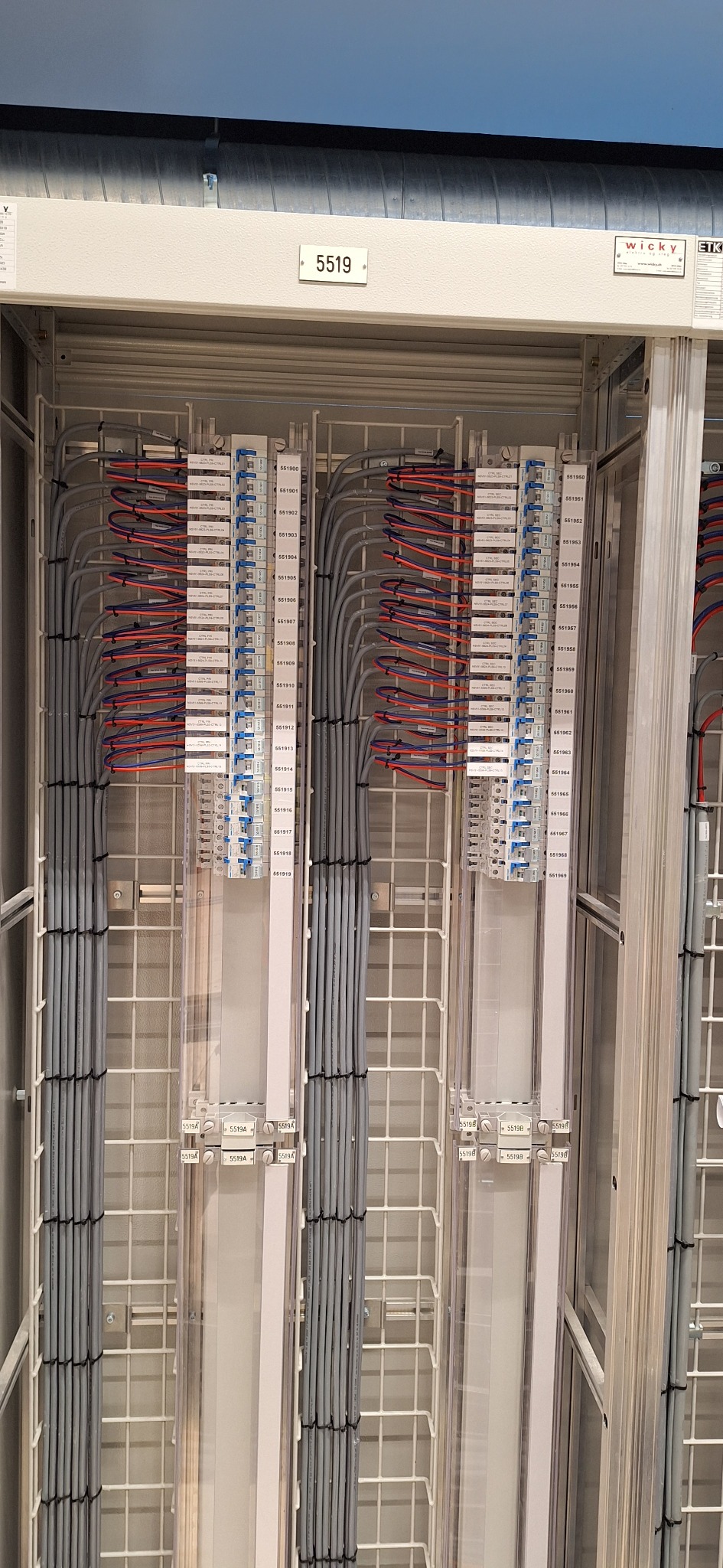 Szafa sterownicza z uporządkowanymi kablami w szarych peszlach, podłączonymi do listew zaciskowych z oznaczeniami numerów. Widoczne detale instalacji elektrycznej.