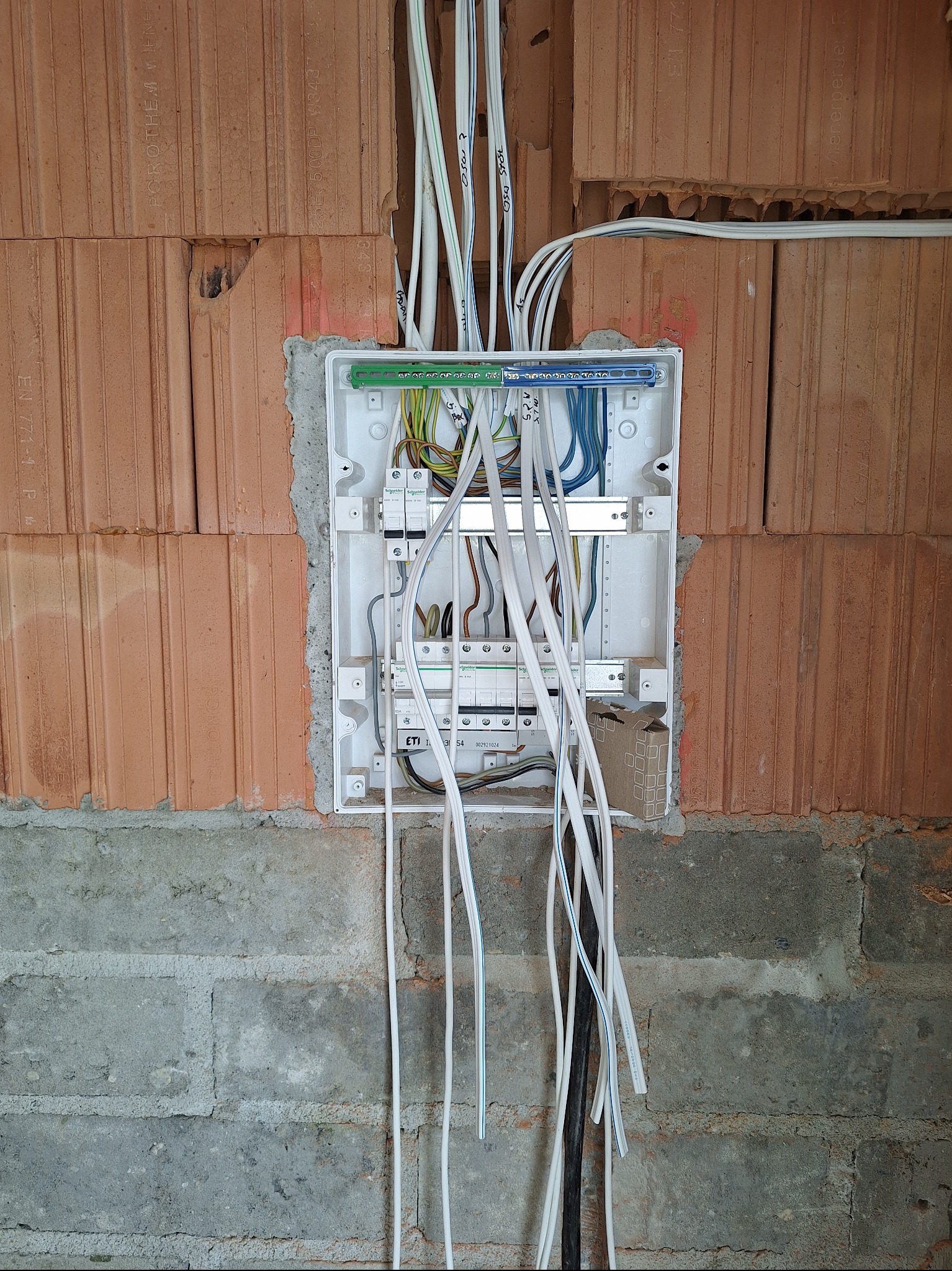 Rozdzielnia elektryczna w trakcie montażu na ścianie z czerwonej cegły. Widoczne kable i zabezpieczenia. Prace instalacyjne w surowym stanie.