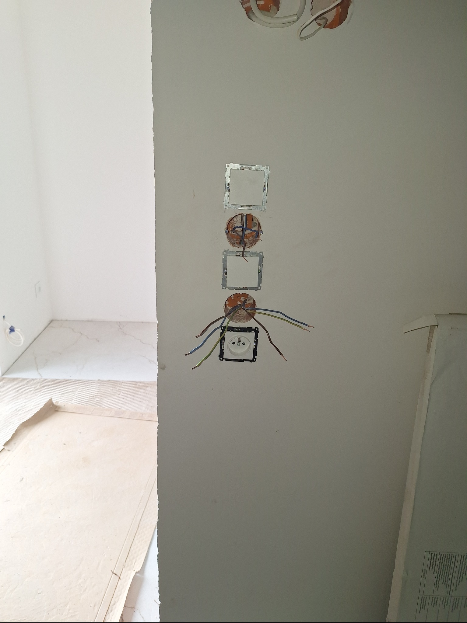 Ściana z przygotowanymi otworami i przewodami pod gniazdka elektryczne i włączniki, widok na wykończoną podłogę w drugim pomieszczeniu. Prace instalacyjne w trakcie remontu.