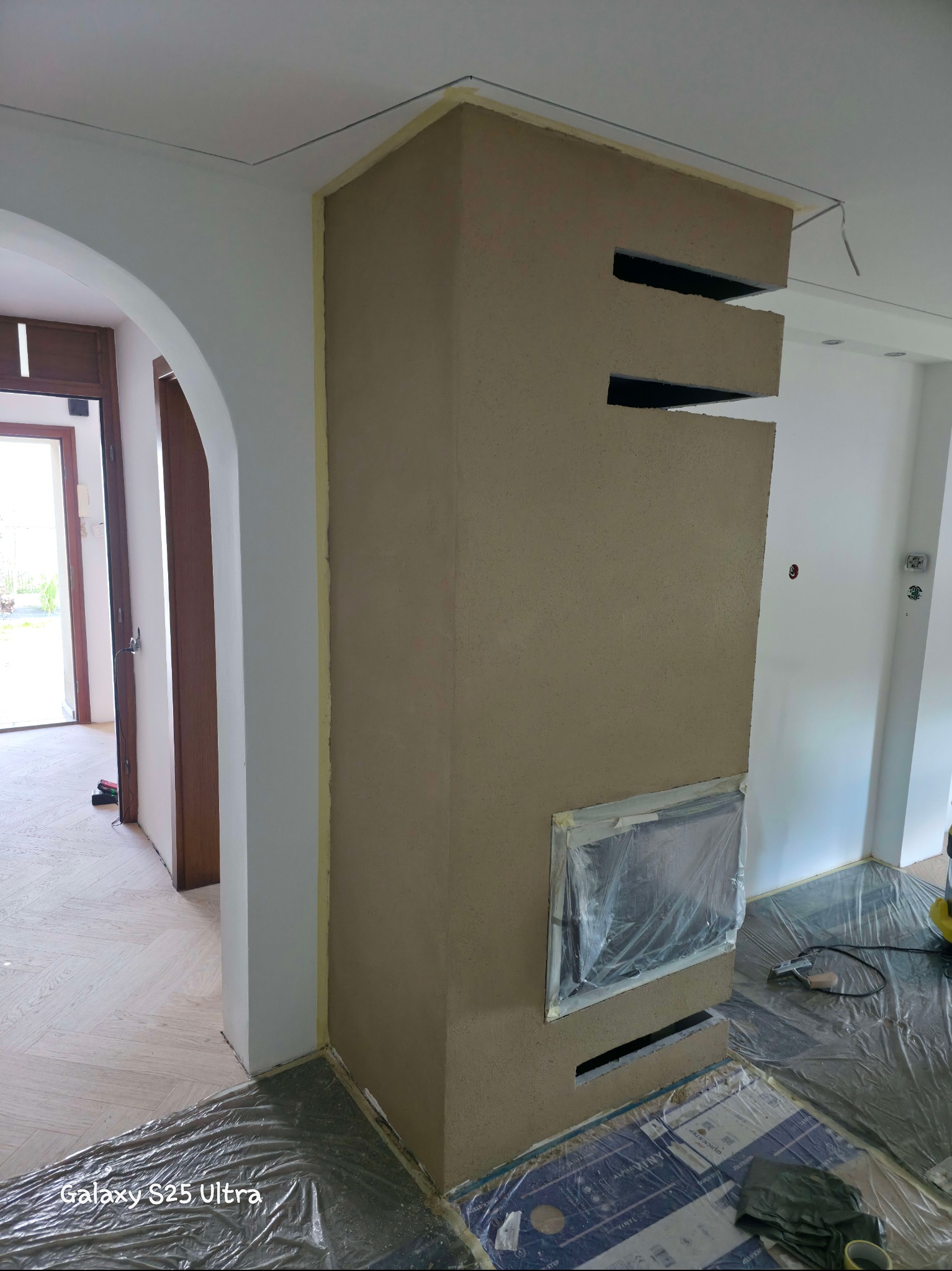 Otynkowany kominek z poziomymi szczelinami, zabezpieczony folią, w trakcie prac wykończeniowych. Widoczne narzędzia i fragment podłogi zabezpieczonej folią malarską.