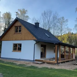 Nowoczesny, parterowy dom z białymi ścianami, drewnianą elewacją szczytową i ciemnym dachem. Widoczny drewniany taras z zadaszeniem i palety z materiałami budowlanymi.