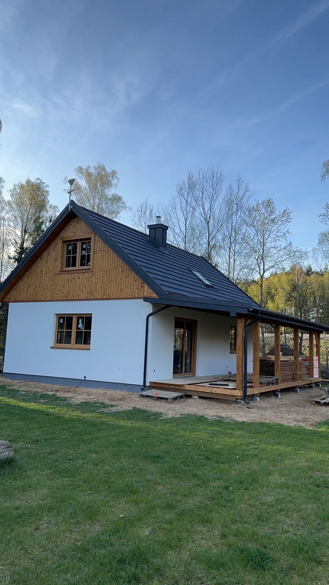 Nowoczesny, parterowy dom z białymi ścianami, drewnianą elewacją szczytową i ciemnym dachem. Widoczny drewniany taras z zadaszeniem i palety z materiałami budowlanymi.