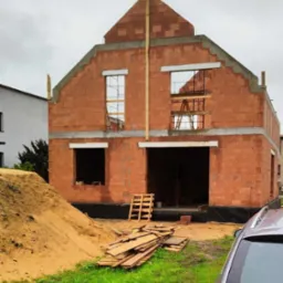 Surowy stan domu z czerwonej cegły, widoczne otwory okienne i drzwiowe, drewniane stemple, paleta i deski na trawniku. Fragment auta w prawym dolnym rogu.