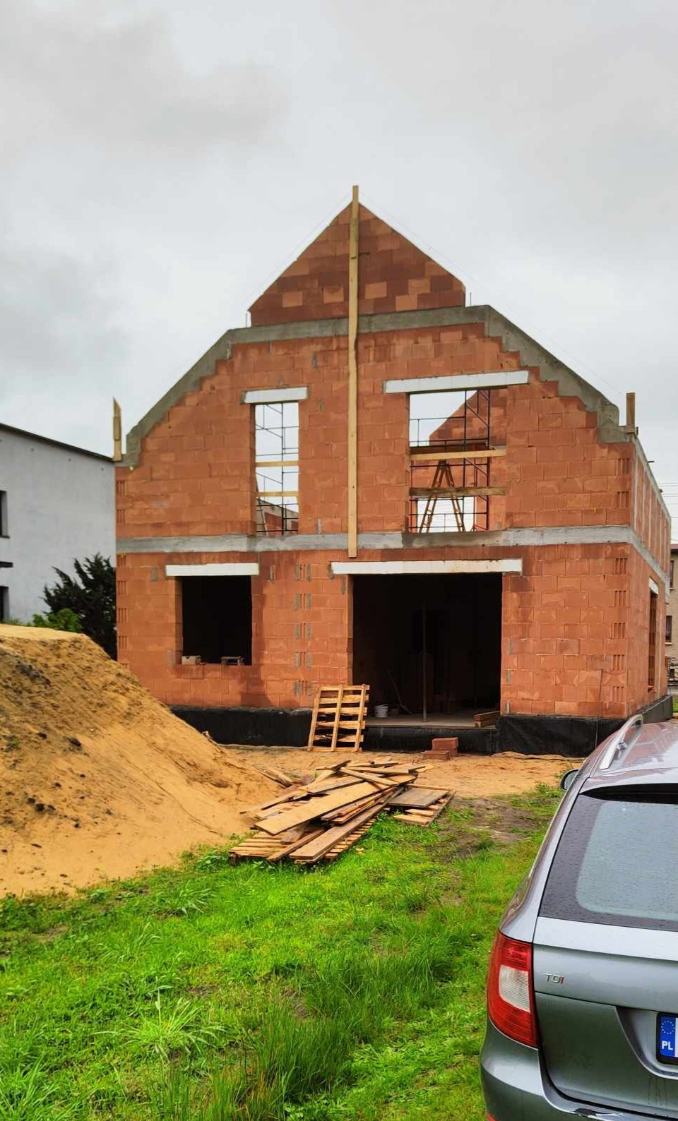 Surowy stan domu z czerwonej cegły, widoczne otwory okienne i drzwiowe, drewniane stemple, paleta i deski na trawniku. Fragment auta w prawym dolnym rogu.