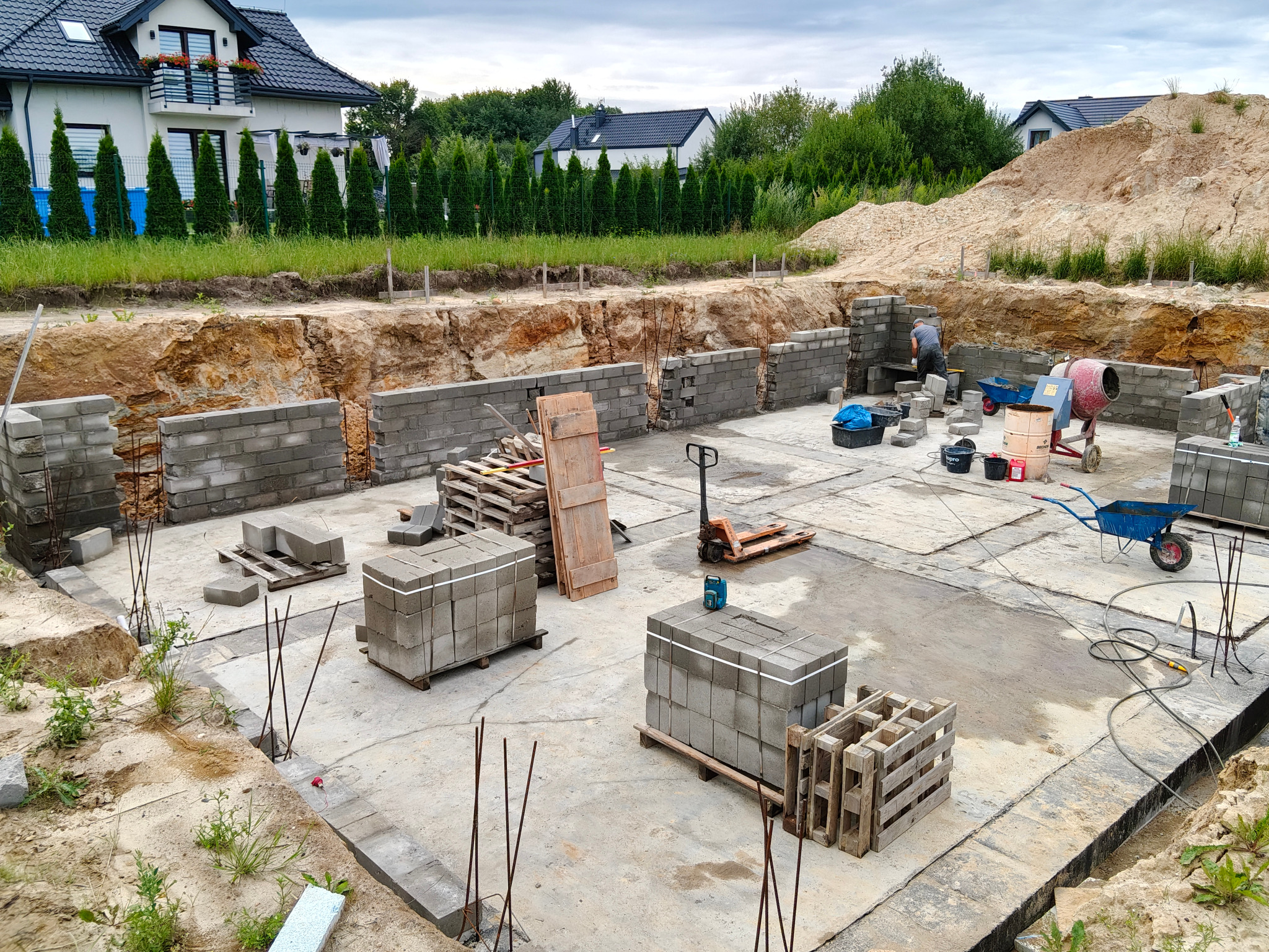 Budowa fundamentów domu jednorodzinnego: częściowo wymurowane ściany z bloczków betonowych, palety z materiałem, pracownik przy pracy, wykop w tle, widok z góry.