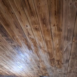 Ogólnobudowlana Mirela Wika - Drewniany sufit z widocznym rysunkiem słojów, ułożony ukośnie. Część sufitu przy ścianie nieukończona, widoczna cegła. Nietypowy kąt ujęcia.