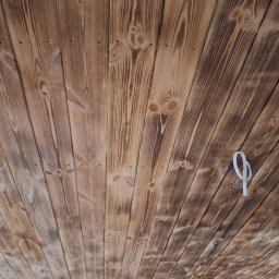 Ogólnobudowlana Mirela Wika - Wykończony drewniany sufit z widocznymi śladami po przypalaniu, ułożony z desek pod kątem. Widoczne mocowania i fragment ściany z cegły.