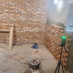 Ogólnobudowlana Mirela Wika - Wnętrze w trakcie prac: częściowo wykończona ściana z cegły, poziomica laserowa na statywie, narzędzia budowlane. Widoczne niedokończone fragmenty ściany.