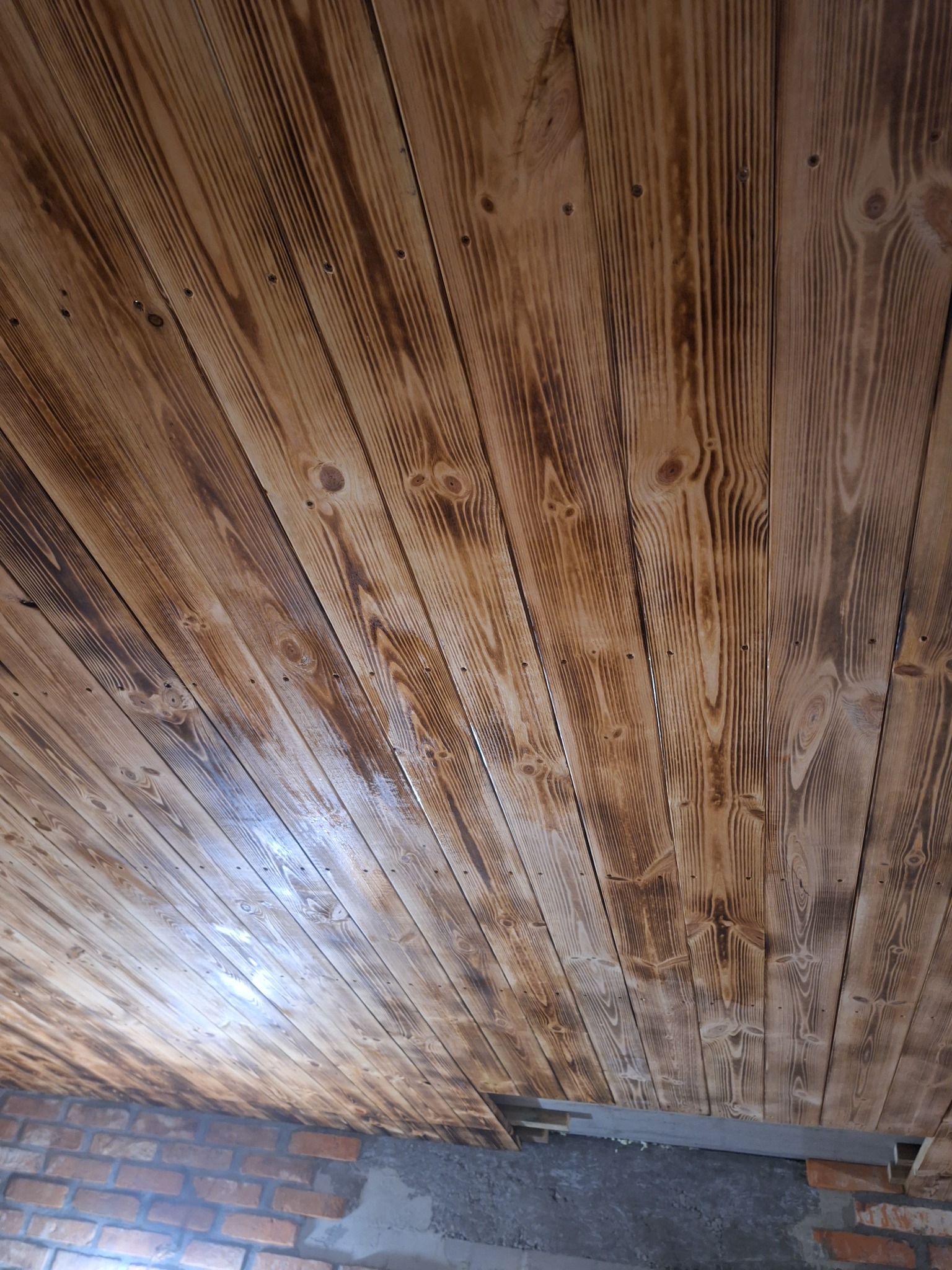 Drewniany sufit z widocznym rysunkiem słojów, ułożony ukośnie. Część sufitu przy ścianie nieukończona, widoczna cegła. Nietypowy kąt ujęcia.