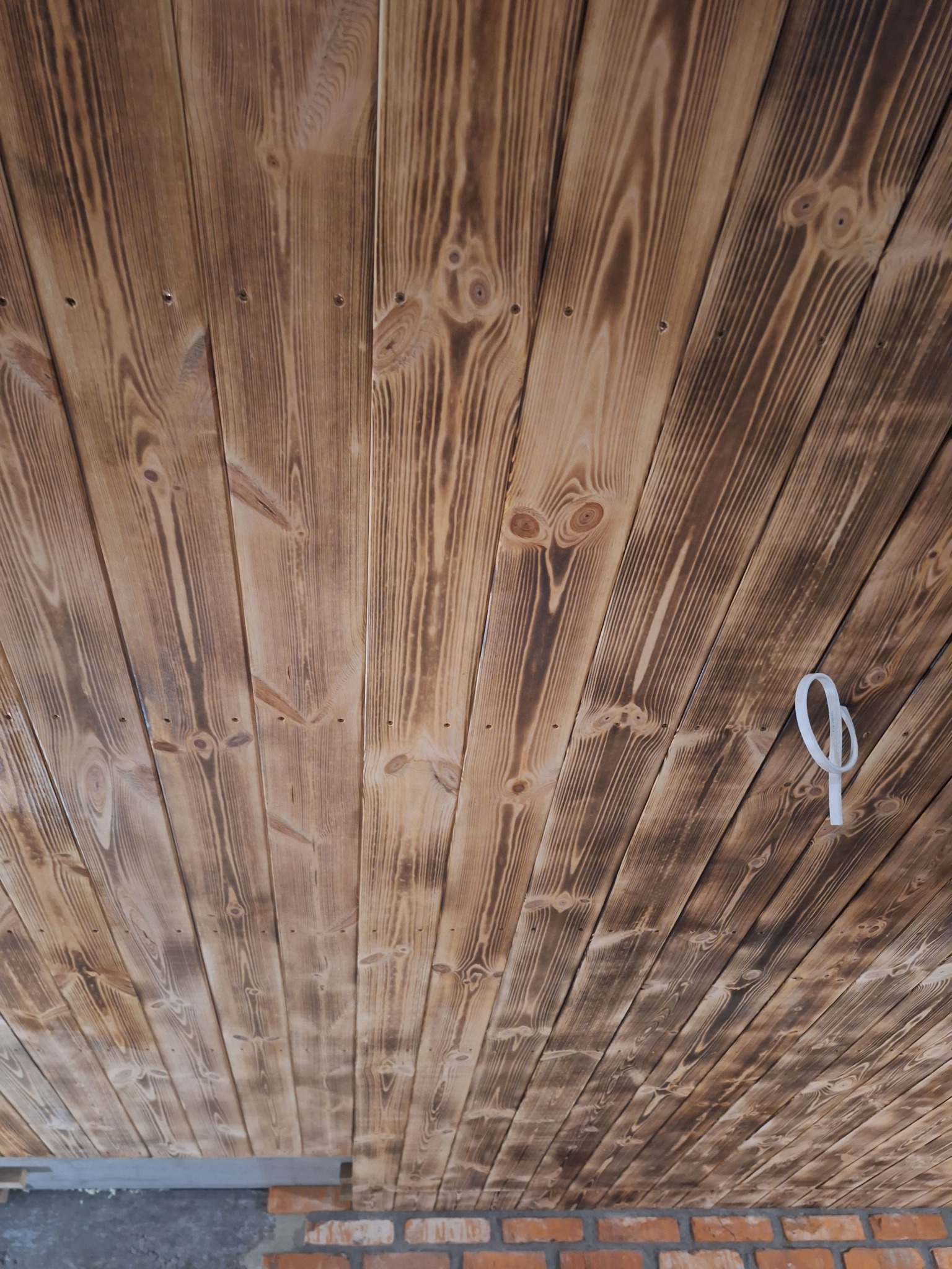 Wykończony drewniany sufit z widocznymi śladami po przypalaniu, ułożony z desek pod kątem. Widoczne mocowania i fragment ściany z cegły.