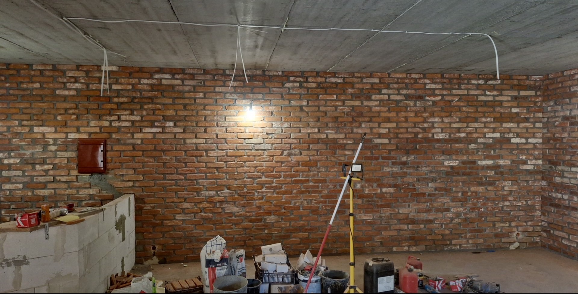 Wnętrze w trakcie remontu: ceglana ściana z widoczną instalacją elektryczną pod sufitem, narzędzia i materiały budowlane na podłodze. Widoczna lampa halogenowa.