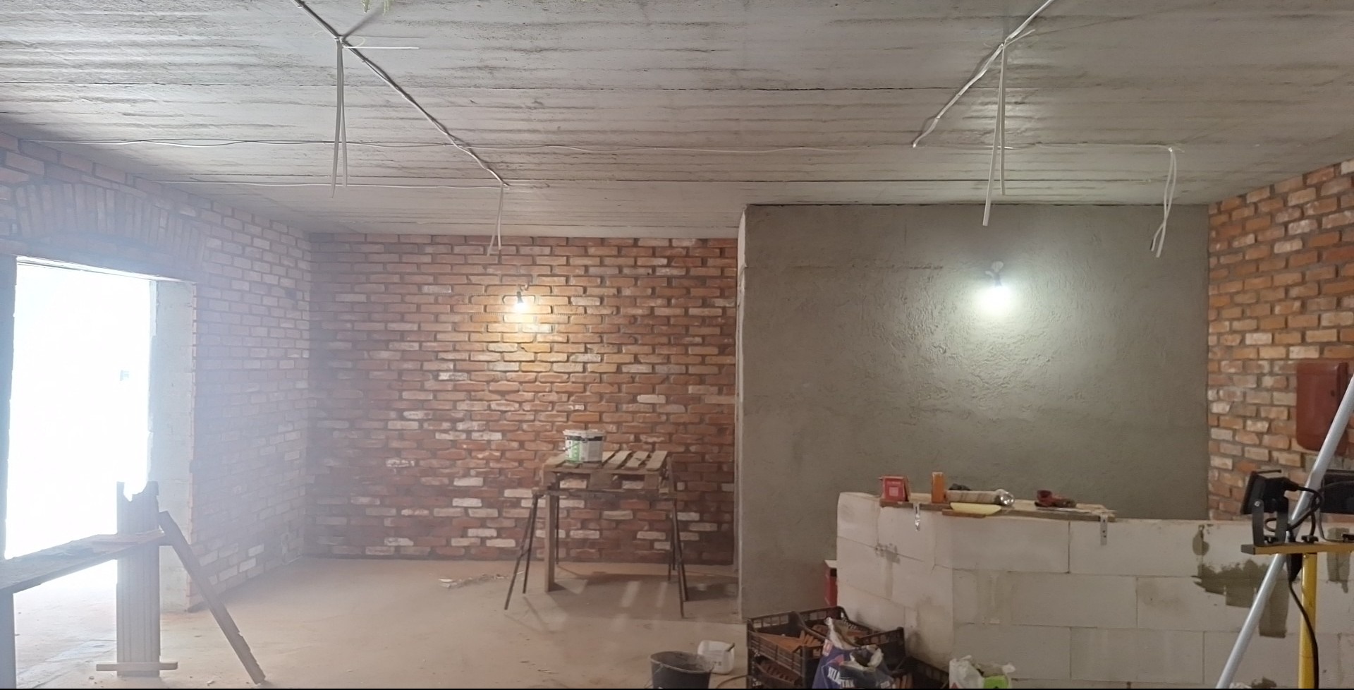 Wnętrze w trakcie remontu: surowy beton na suficie, fragment ściany z cegły, oświetlenie budowlane i narzędzia na tle ściany pokrytej świeżym tynkiem.