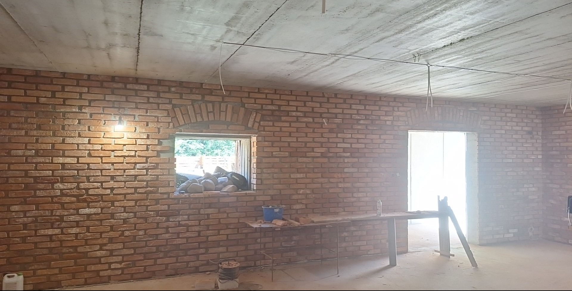 Murowana ściana z cegły z łukowym oknem i otworem drzwiowym. Widoczne narzędzia i materiały budowlane. Surowy betonowy sufit z instalacją elektryczną.