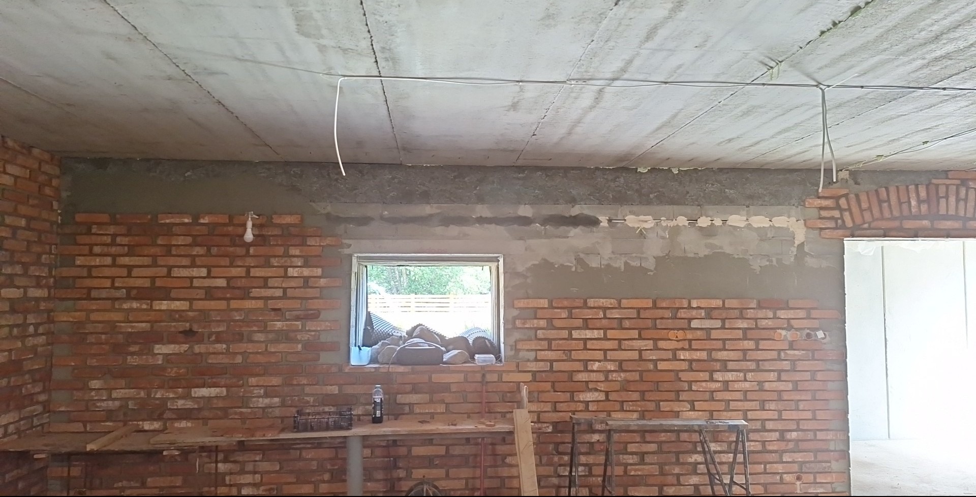 Ceglana ściana w trakcie tworzenia, widoczne zaprawy i niedokończone fragmenty. Okno z widokiem na zieleń w tle. Na suficie widoczne kable i surowy beton.
