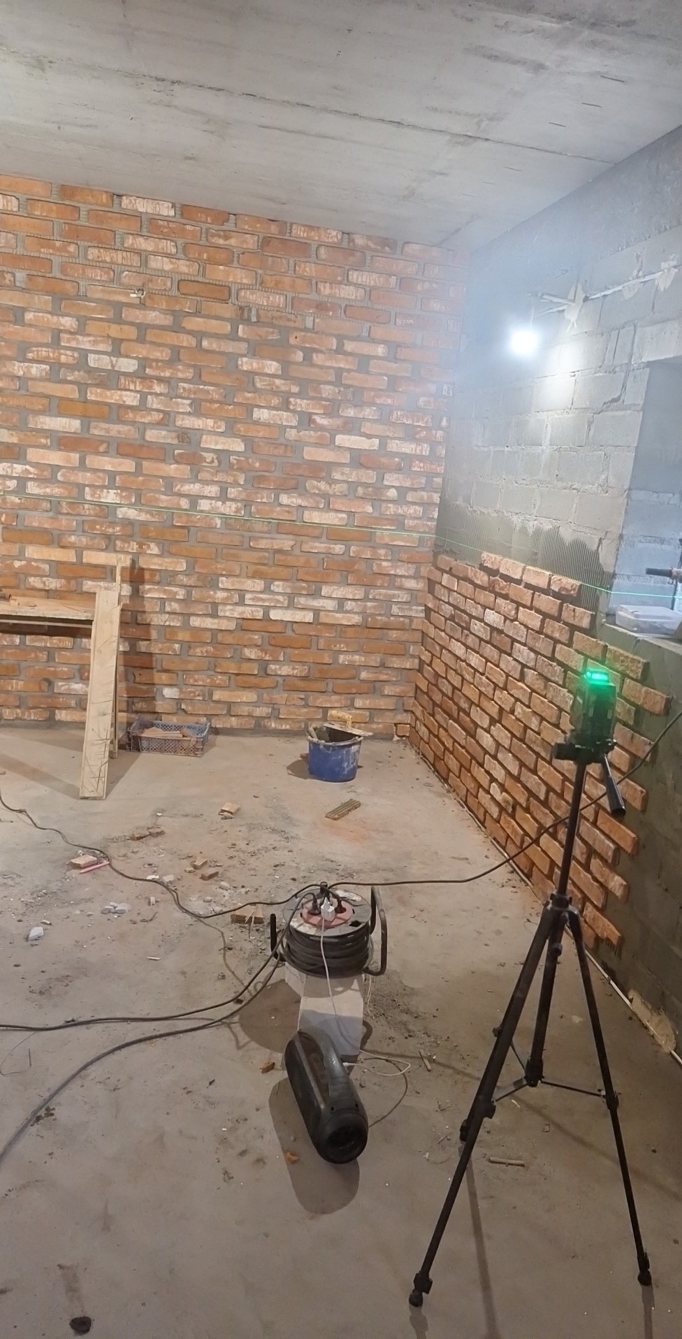 Wnętrze w trakcie prac: częściowo wykończona ściana z cegły, poziomica laserowa na statywie, narzędzia budowlane. Widoczne niedokończone fragmenty ściany.