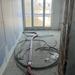 Instalacja ogrzewania podłogowego w surowym stanie: czerwone rury grzewcze ułożone na szarym betonie w pomieszczeniu z oknem i ocieplonymi ścianami.