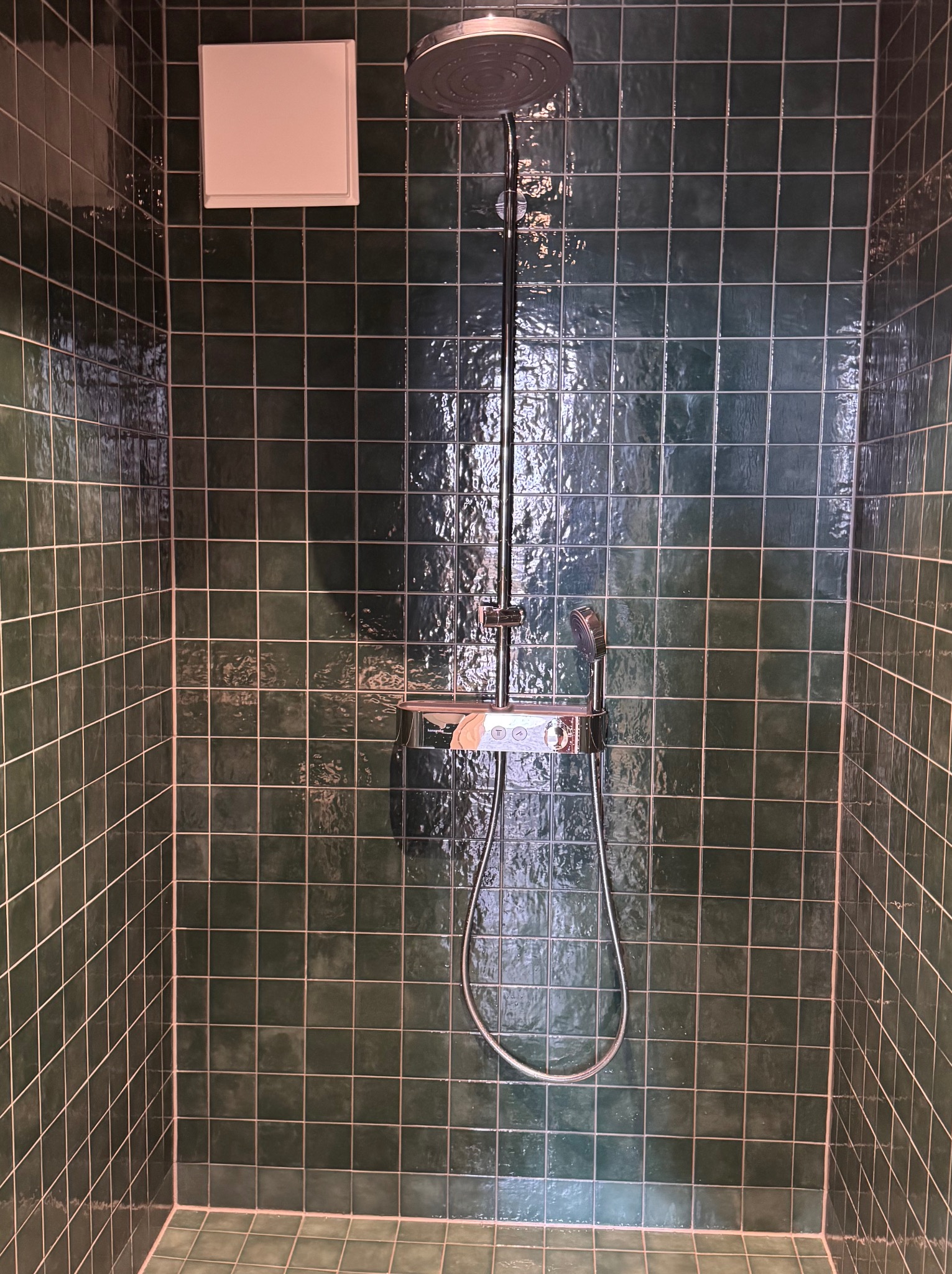 Chromowana bateria prysznicowa z deszczownicą i słuchawką na ścianie wyłożonej ciemnozieloną, kwadratową glazurą w nowoczesnej kabinie prysznicowej.