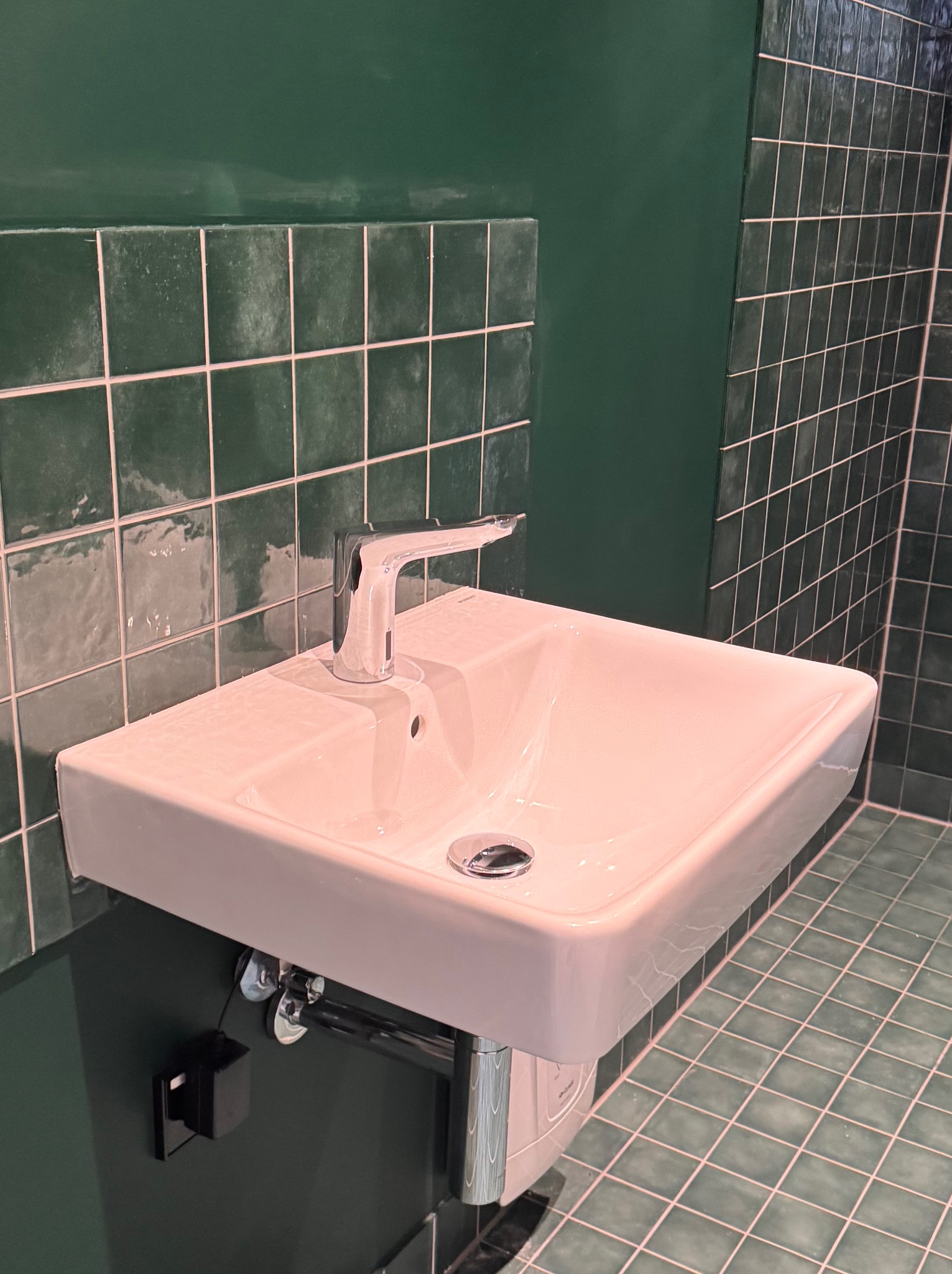 Biała umywalka z chromowaną baterią na tle ściany i podłogi w zielonej, kwadratowej glazurze. Umywalka zamontowana na ścianie, widoczne elementy instalacji.