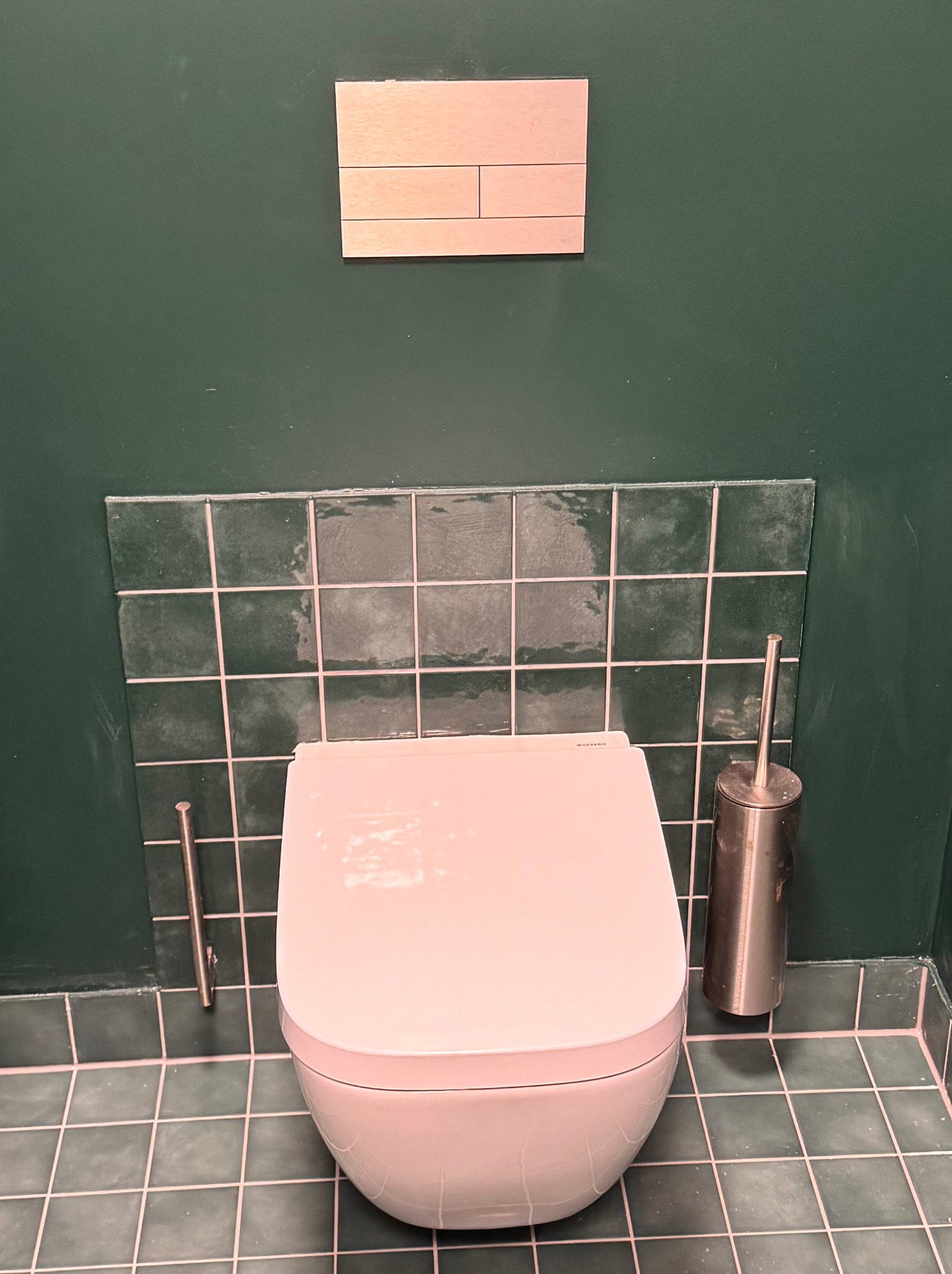 Nowoczesna toaleta podwieszana w łazience z zielonymi płytkami i ukrytą spłuczką. Widoczny uchwyt na papier toaletowy i szczotka do WC.