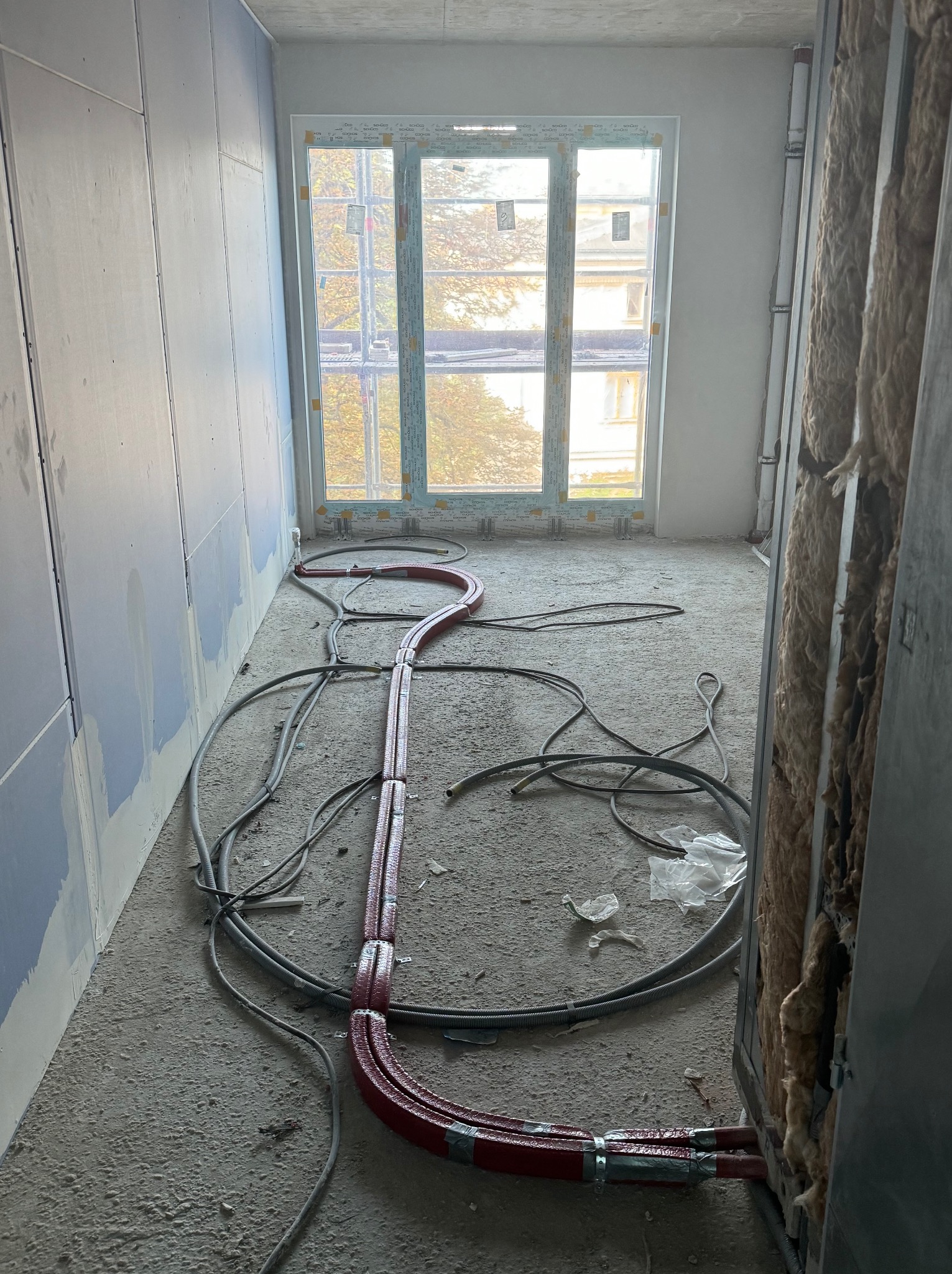 Instalacja ogrzewania podłogowego w surowym stanie: czerwone rury grzewcze ułożone na szarym betonie w pomieszczeniu z oknem i ocieplonymi ścianami.