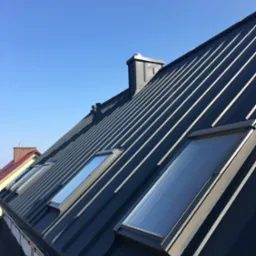 Ciemnoszary dach z blachy na rąbek stojący, z oknami dachowymi i kominem, widziany pod kątem na tle błękitnego nieba. Widoczne fragmenty innych dachów.