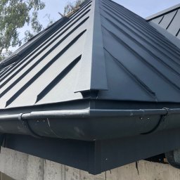 Dekarz Sp z o.o. - Szary dach z blachy na rąbek stojący, widok z dołu, z fragmentem rynny i surowego muru. Widoczne łączenia blachy i elementy mocujące rynnę.