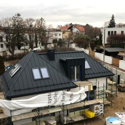 Dekarz Sp z o.o. - Dom z czarnym dachem z blachy na rąbek stojący, widok z góry. Na dachu okna połaciowe. W tle zabudowa miejska i drzewa. Częściowo widoczne rusztowania.