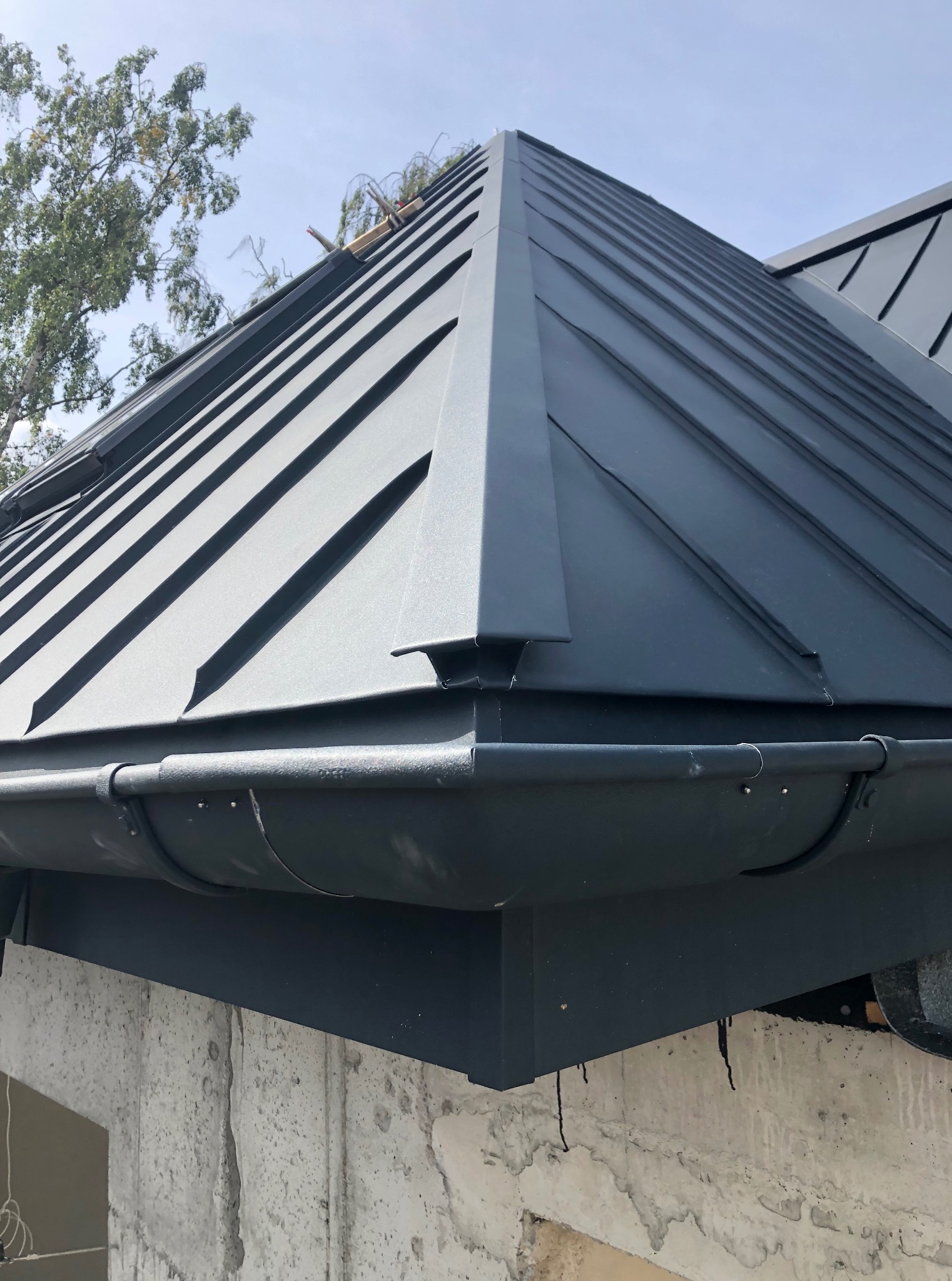 Szary dach z blachy na rąbek stojący, widok z dołu, z fragmentem rynny i surowego muru. Widoczne łączenia blachy i elementy mocujące rynnę.