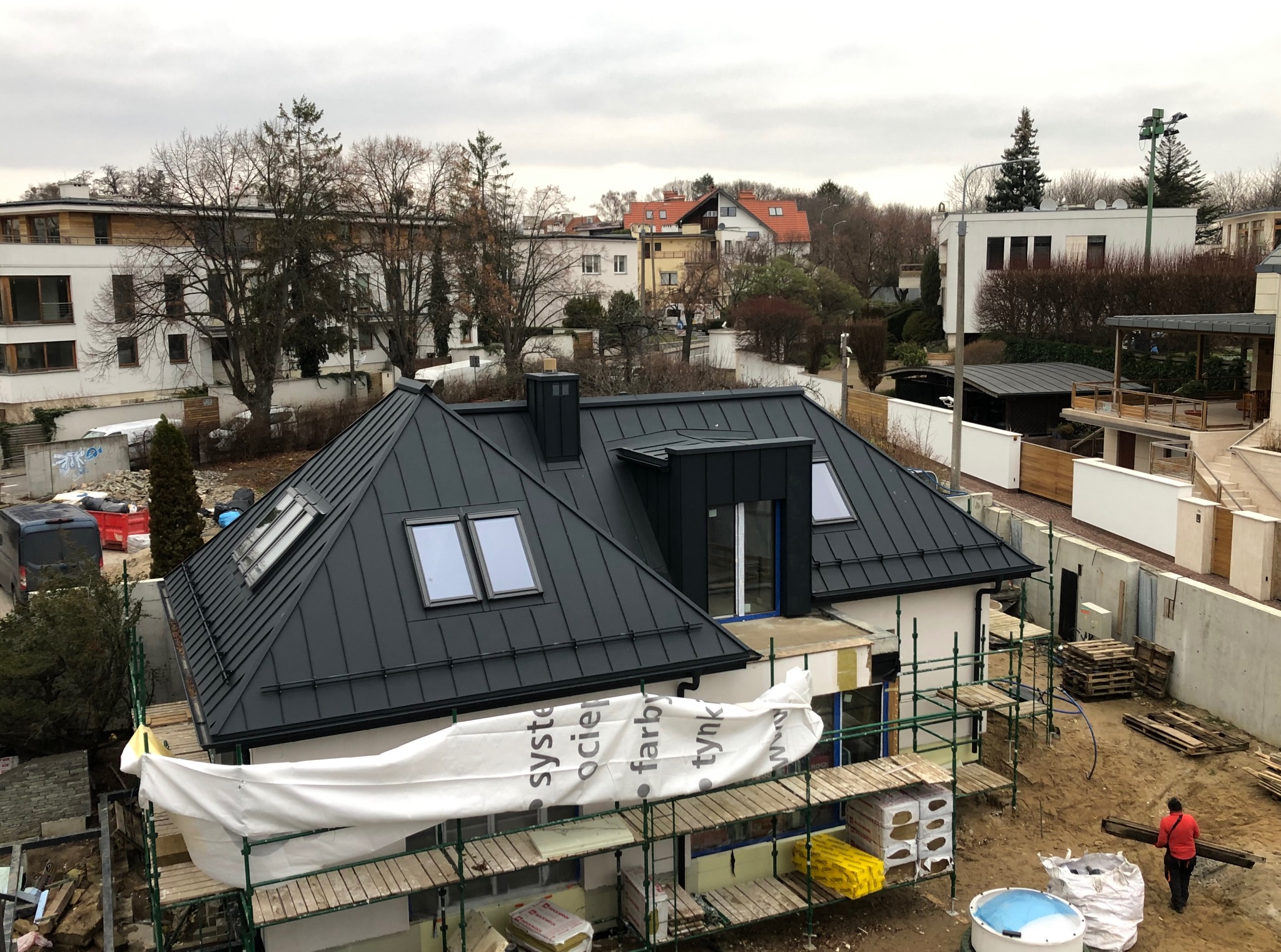 Dom z czarnym dachem z blachy na rąbek stojący, widok z góry. Na dachu okna połaciowe. W tle zabudowa miejska i drzewa. Częściowo widoczne rusztowania.