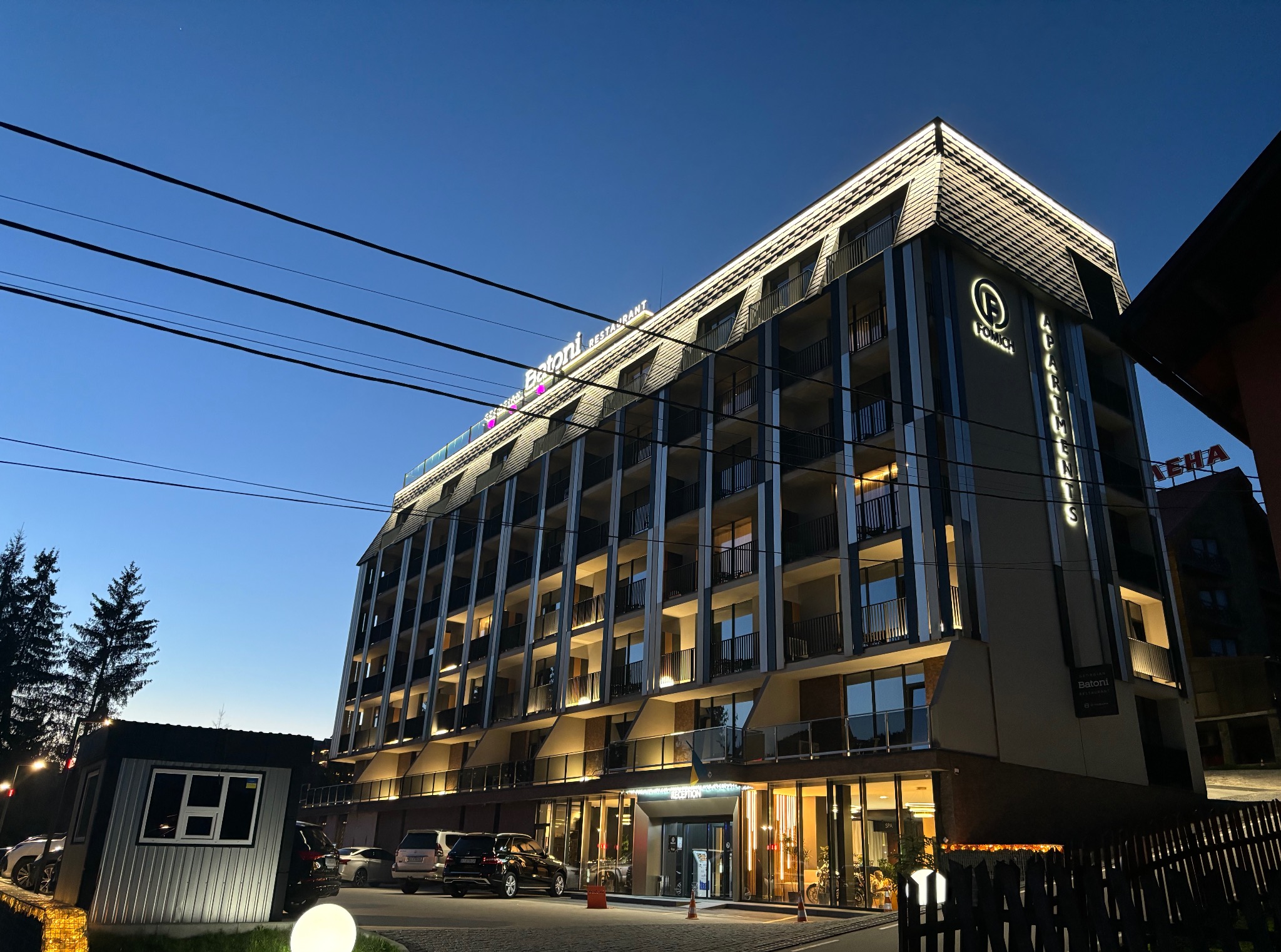 Elewacja budynku apartamentowego z podświetlonymi napisami 'Batoni Restaurant' i 'Fomich Apartments' o zmierzchu. Widoczne linie energetyczne.