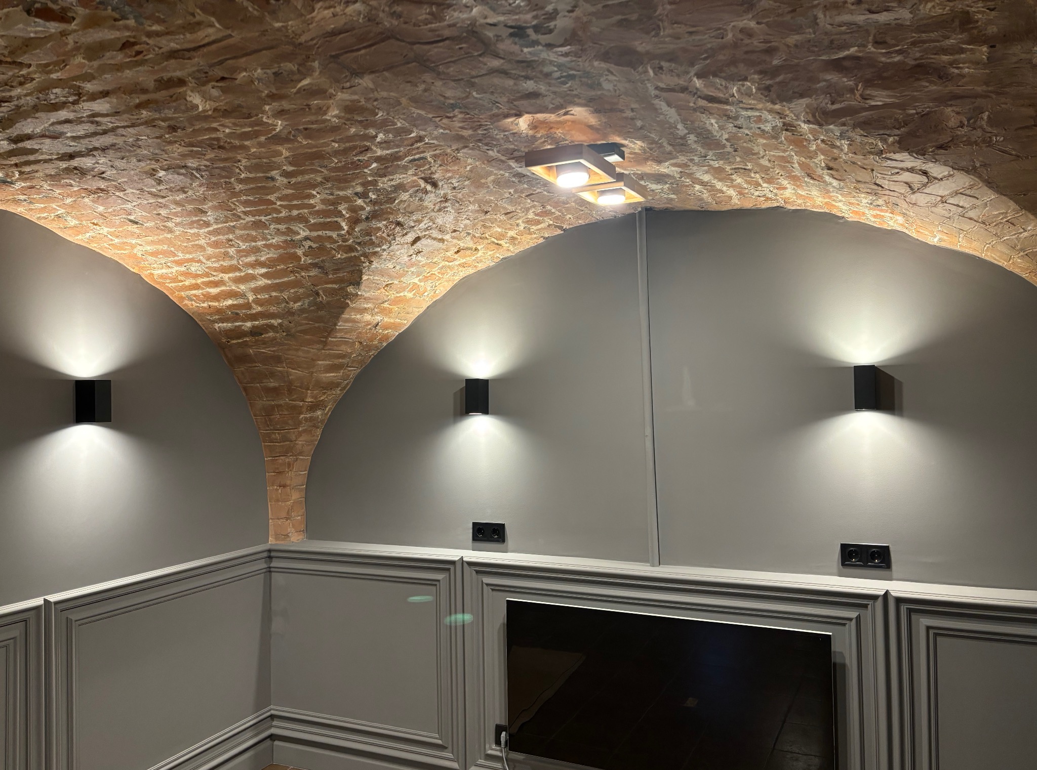 Oświetlenie ścienne i sufitowe w pomieszczeniu z ceglanym sklepieniem. Czarne, kwadratowe lampy emitują jasne światło. Widoczne gniazdka elektryczne i dekoracyjne panele ścienne.