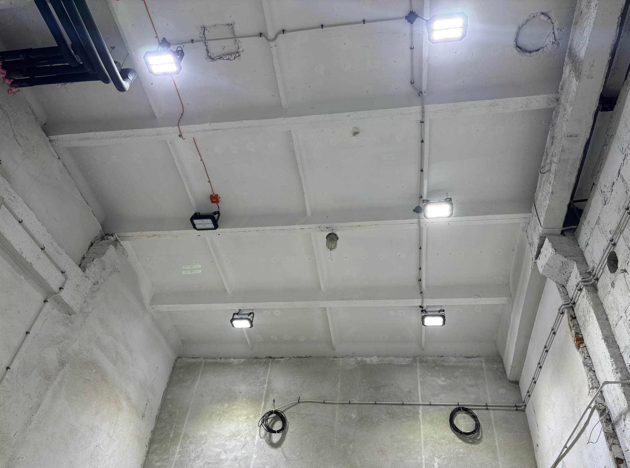 Białe, surowe wnętrze z widocznymi instalacjami elektrycznymi i oświetleniem LED. Kable prowadzone po suficie i ścianach, surowy tynk. Widoczne rury i elementy konstrukcyjne.