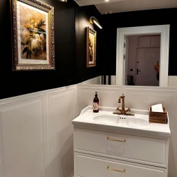 Ale Zarys Aleksandra Onak - Elegancka łazienka w czerni i bieli: umywalka z szafką, złota armatura, obrazy podświetlone kinkietami, lustro i czarno-biała podłoga w romby.