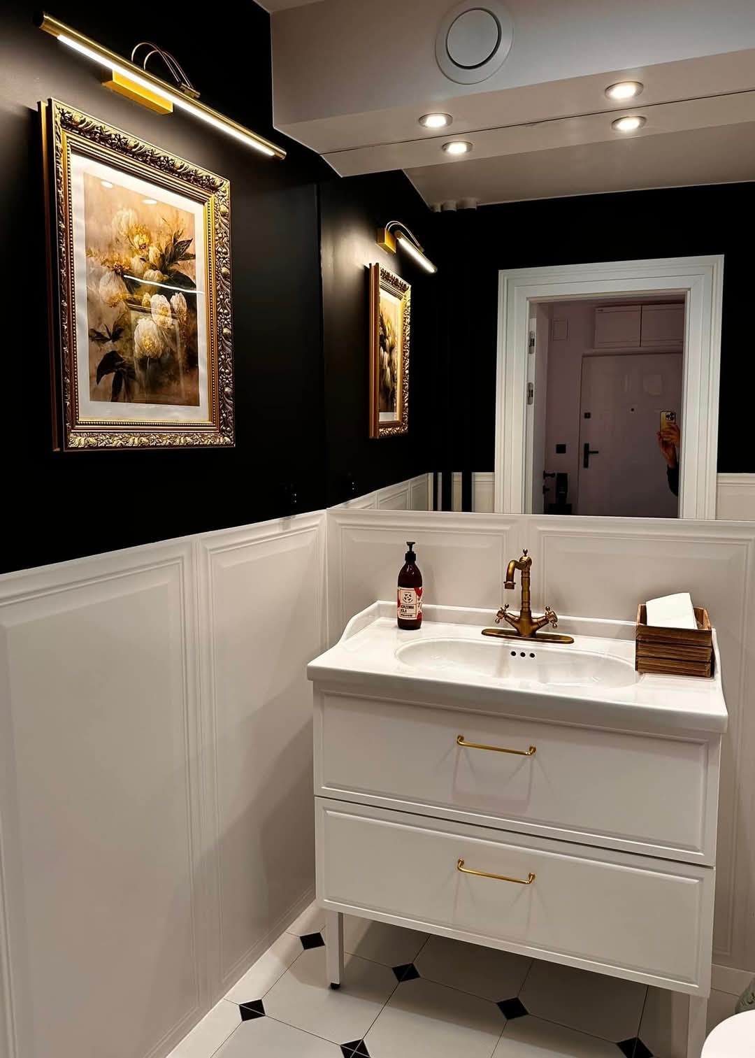 Elegancka łazienka w czerni i bieli: umywalka z szafką, złota armatura, obrazy podświetlone kinkietami, lustro i czarno-biała podłoga w romby.