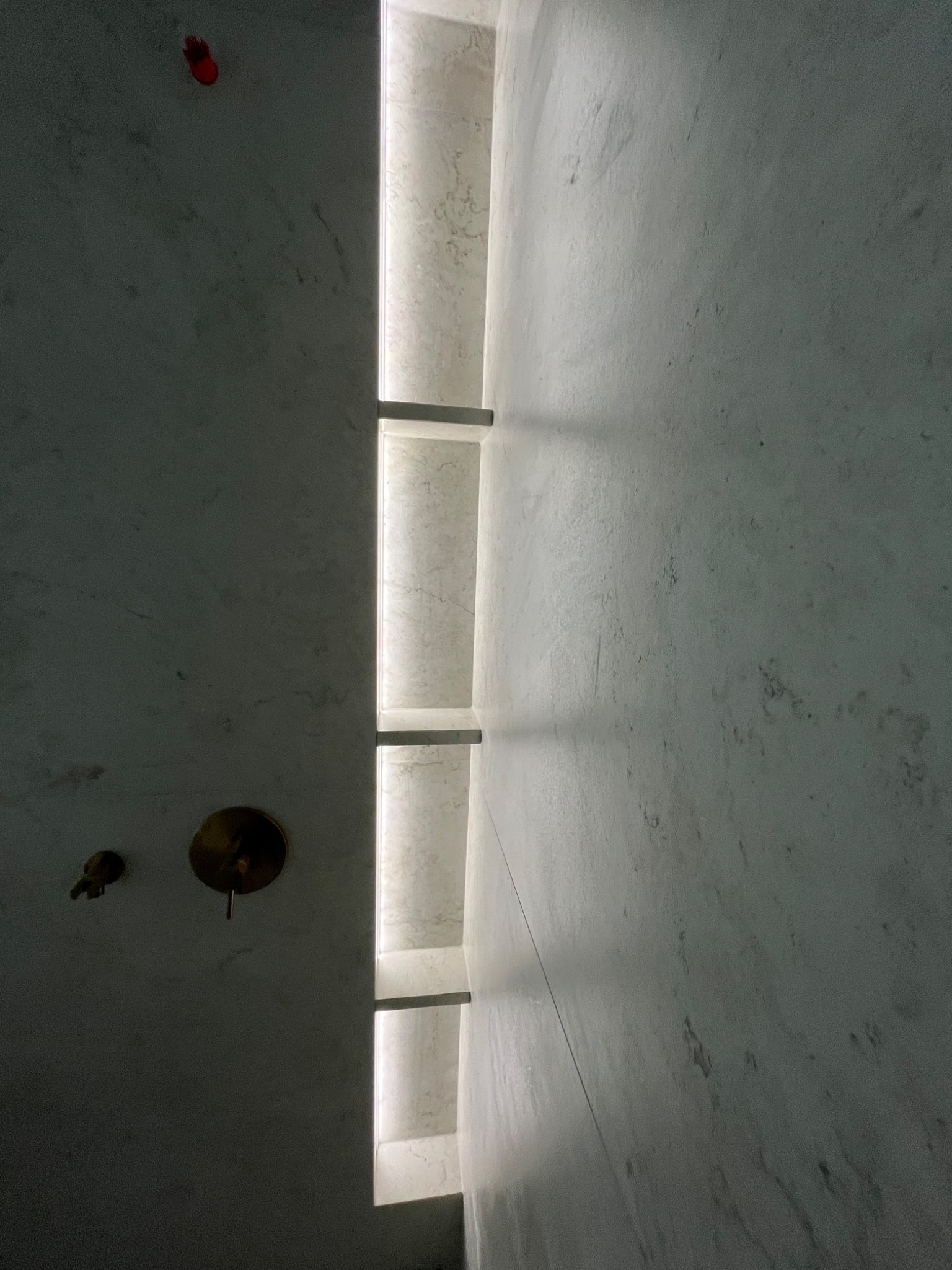 Minimalistyczna łazienka z marmurowymi ścianami, złotą armaturą i pionowym oświetleniem wnękowym. Czerwony element dekoracyjny na ścianie dodaje unikalnego charakteru.