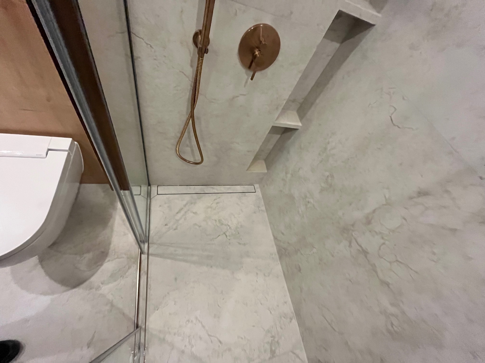 Nowoczesna kabina prysznicowa z marmurowymi płytkami, złotą armaturą i wnękami w ścianie. Widoczny odpływ liniowy i fragment toalety. Minimalistyczny design.